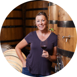 Kimberlee in front of wine barrels, holding a glass of red wine.