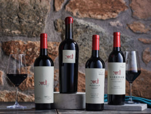 Four Markham red wine bottles in front of a stone wall, with two glasses of red wine. The Holiday collection.