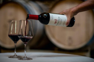 Wine barrels in the background, person pouring red wine into two glasses.