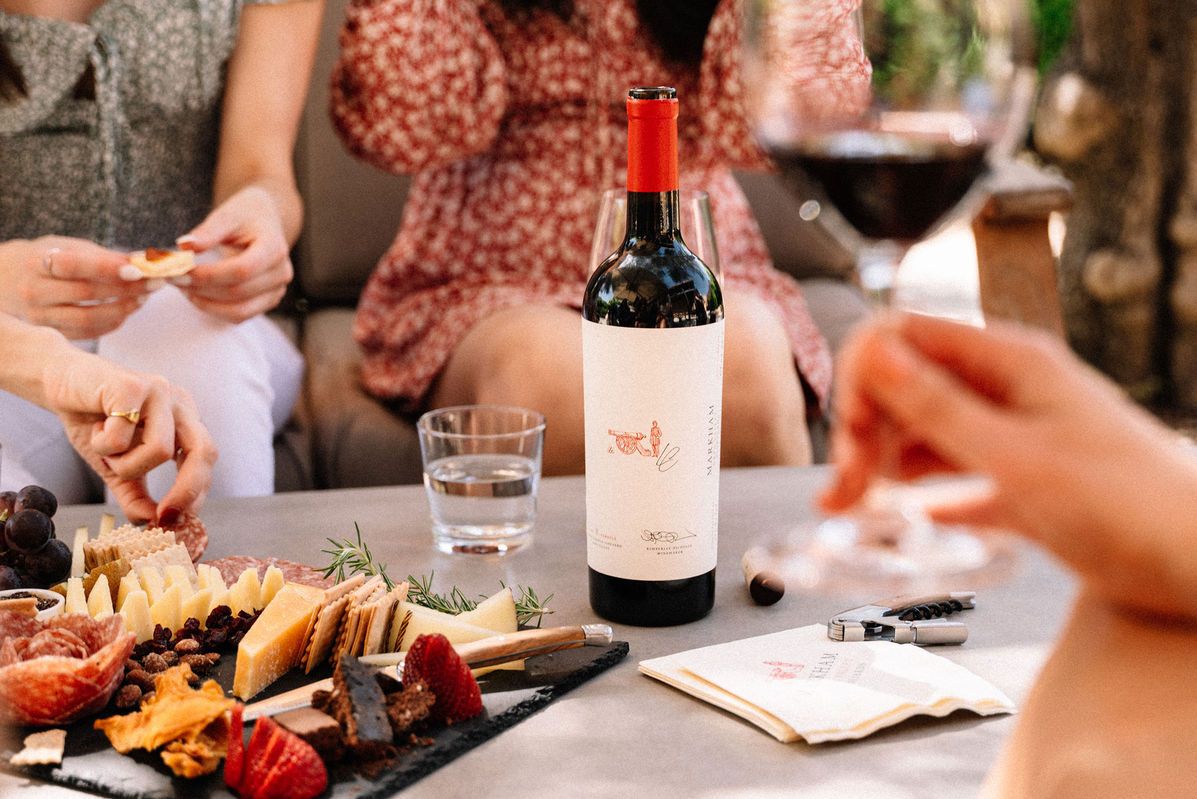 People around a low table with cheese & charcuterie and bottle of markham wine