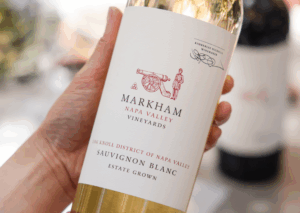 Person holding bottle of Markham wine with a table with wine glasses in the background