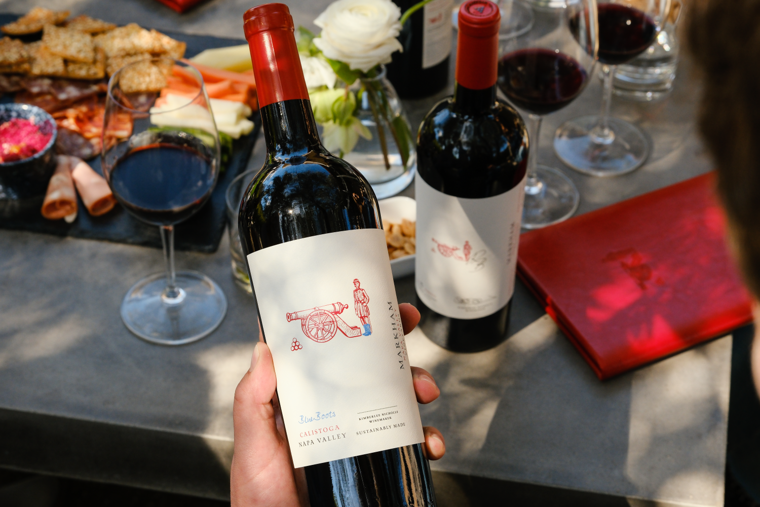 A hand holding a bottles of Blue Boots Red Blend with a table with food pairings in the background