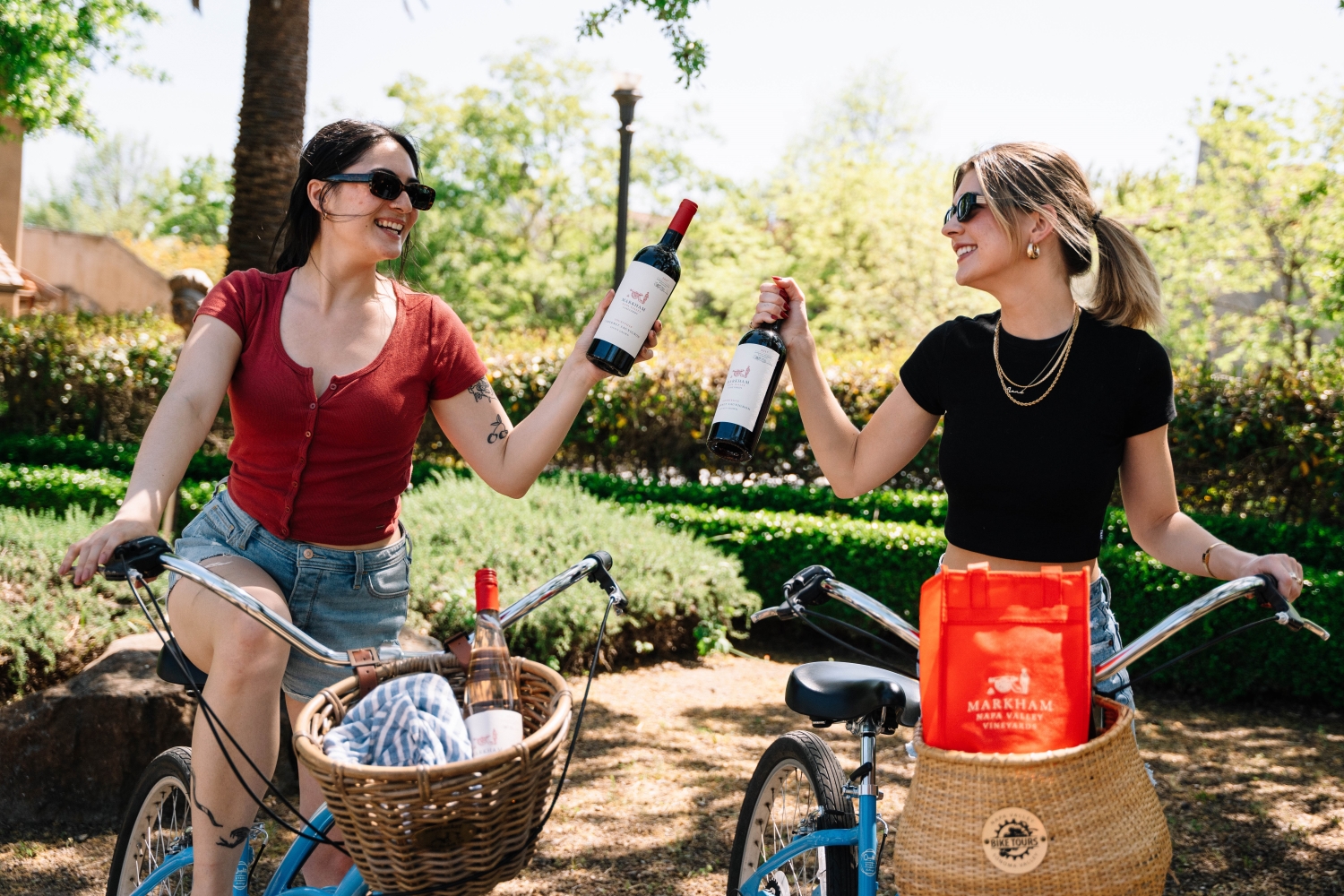 Two people on bikes holding up bottles of Markham wine
